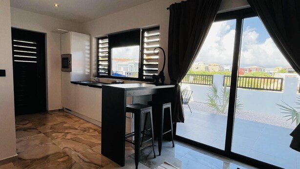 A modern kitchen with a built-in oven, a dining area with a bar stool, and a large sliding glass door leading to a balcony with a view of the city.