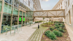 Un edificio moderno con amplios ventanales, un área de estar exterior con sillas y mesas, y un jardín ajardinado con plantas.