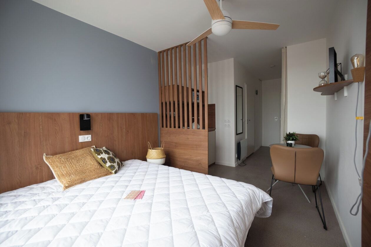 A hotel room with a bed, a ceiling fan, a wooden partition, a small table with a chair, and a shelf with decorative items.