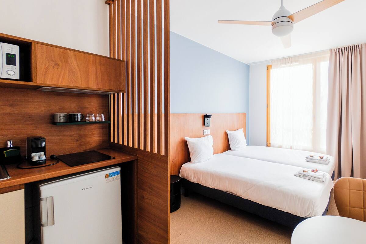 A hotel room with a kitchenette, two beds, a ceiling fan, and a large window with curtains.