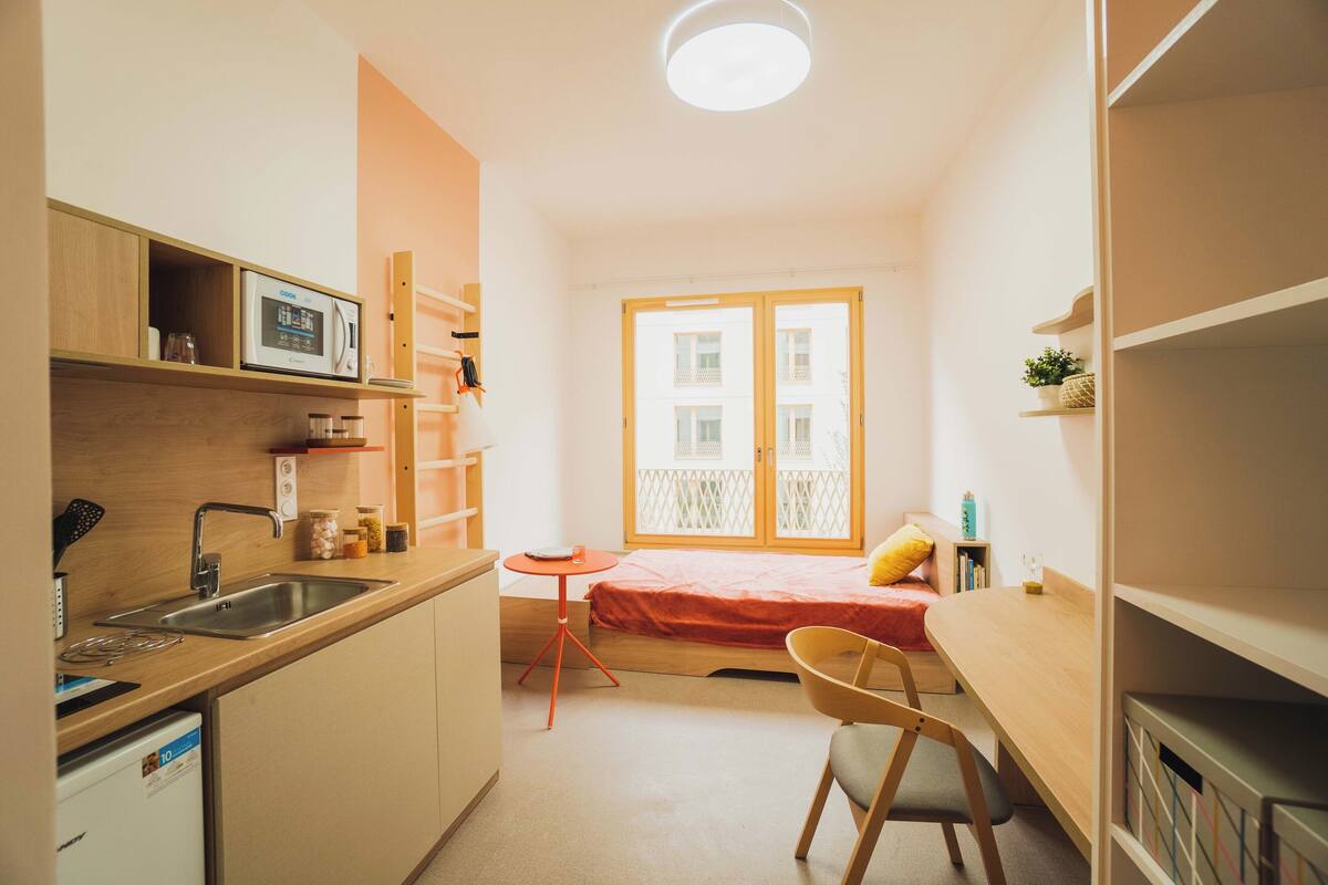 A compact kitchen with a microwave, sink, and a small table. There is a bed with a red blanket and a chair.