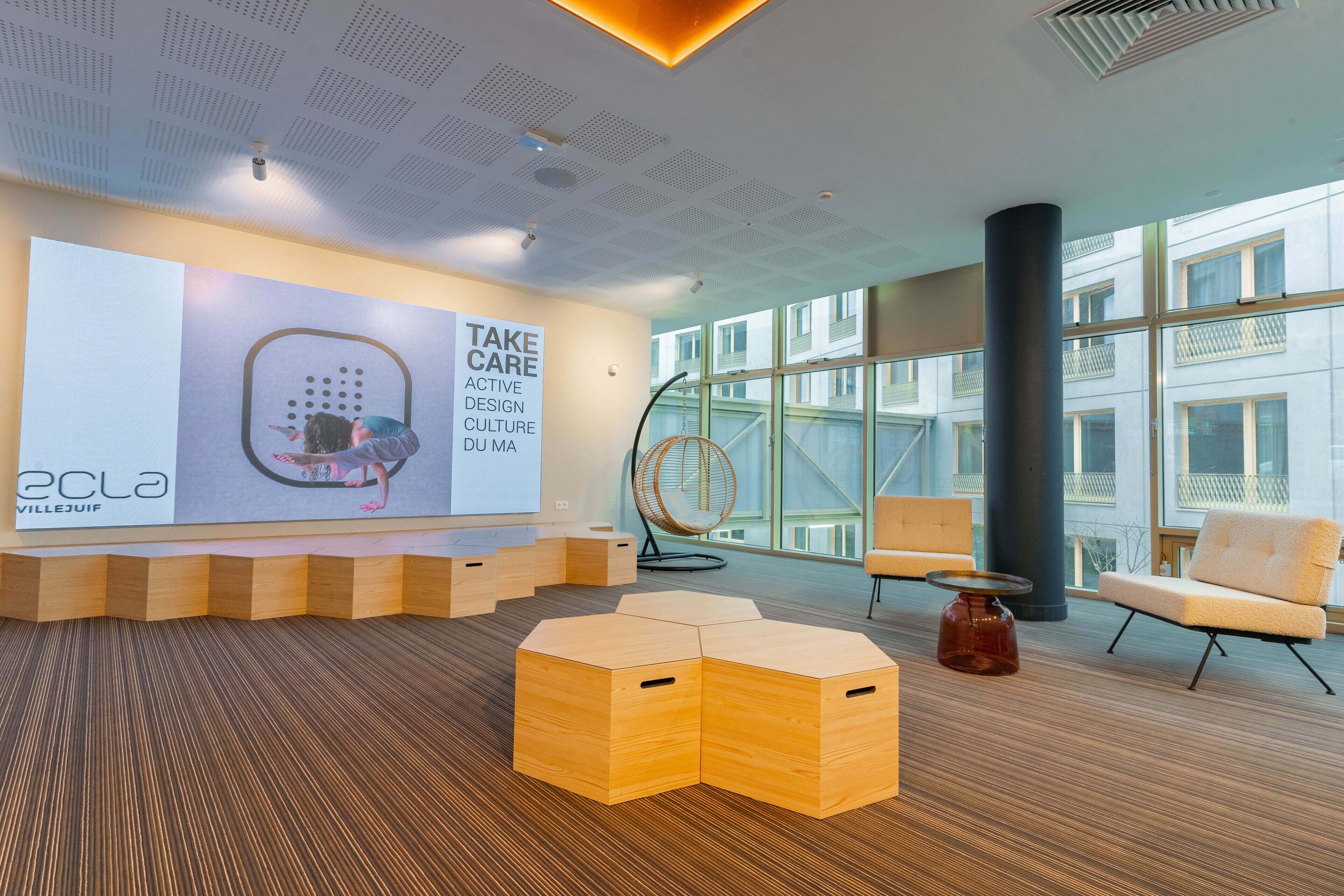 A modern lobby with a large digital display, wooden seating, and large windows offering views of the building.