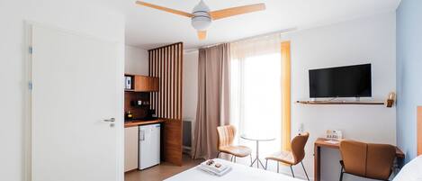 A hotel room with a bed, a desk, a TV, a ceiling fan, and a kitchenette.