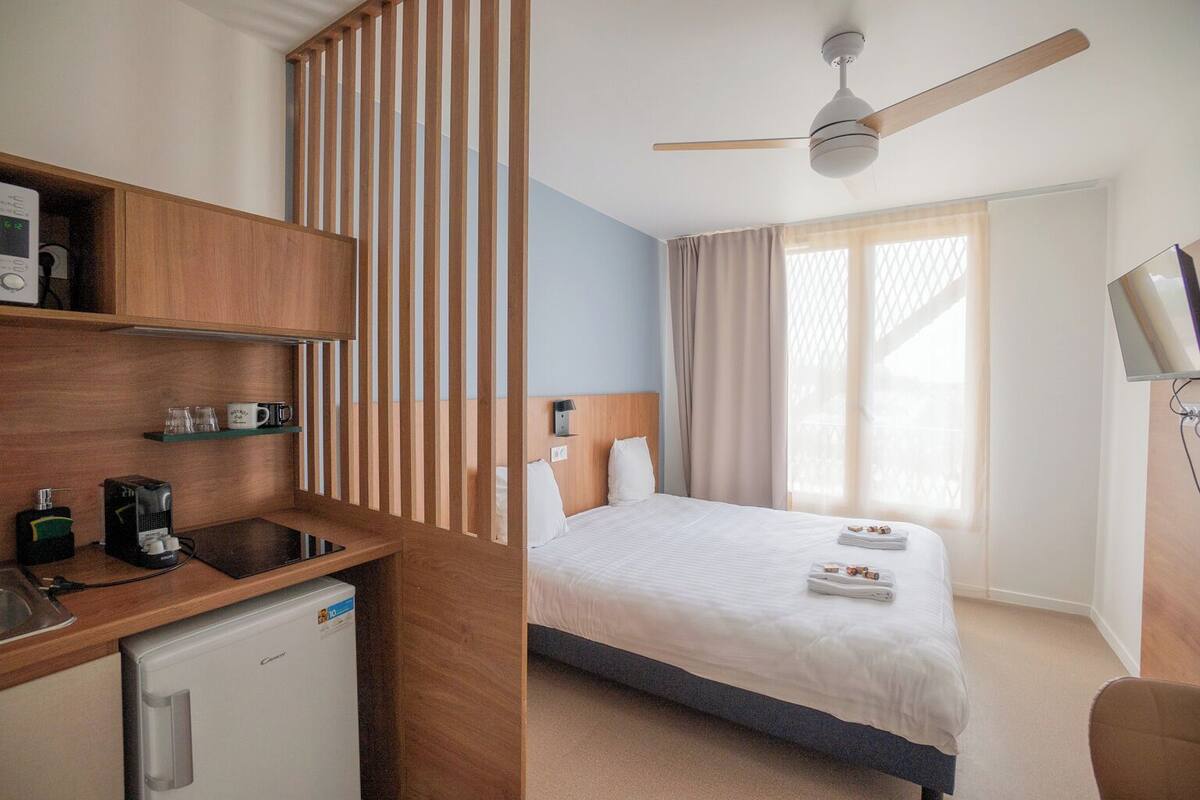 A hotel room with a bed, a desk with a coffee maker, a refrigerator, a TV, and a ceiling fan.
