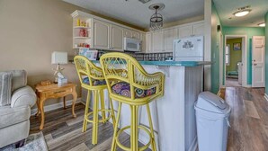 A compact kitchen with white cabinets, a microwave, and a refrigerator. There are two yellow bar stools with patterned cushions. A white trash bin is visible to the right. - Villa Capriani 204B - Oceanfront - Pools - Hot Tubs - Restaurant (North Topsail Beach)