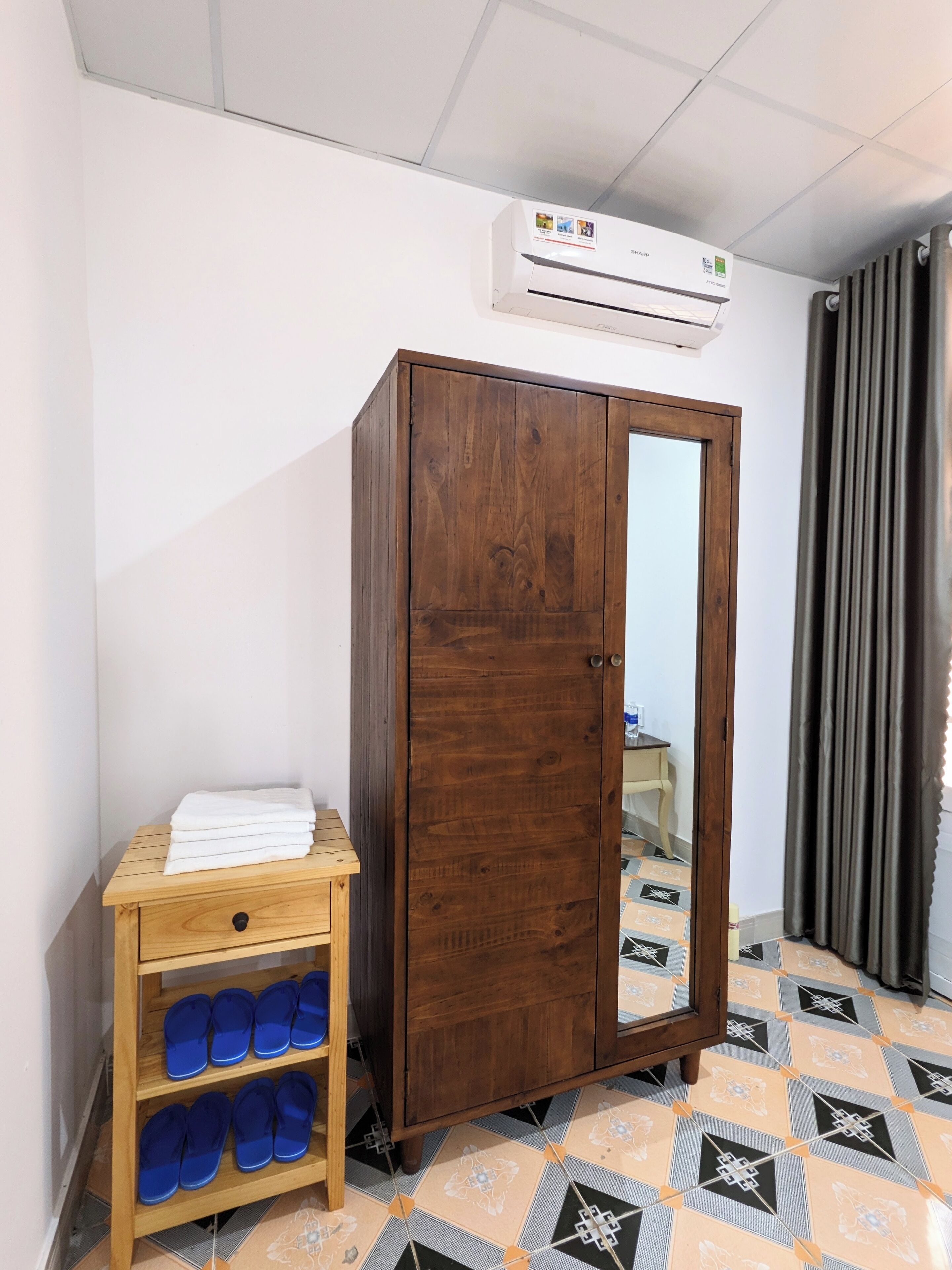 Una habitación con un armario de madera, un pequeño estante con toallas dobladas y pantuflas azules, suelo de baldosas y un equipo de aire acondicionado en la pared.