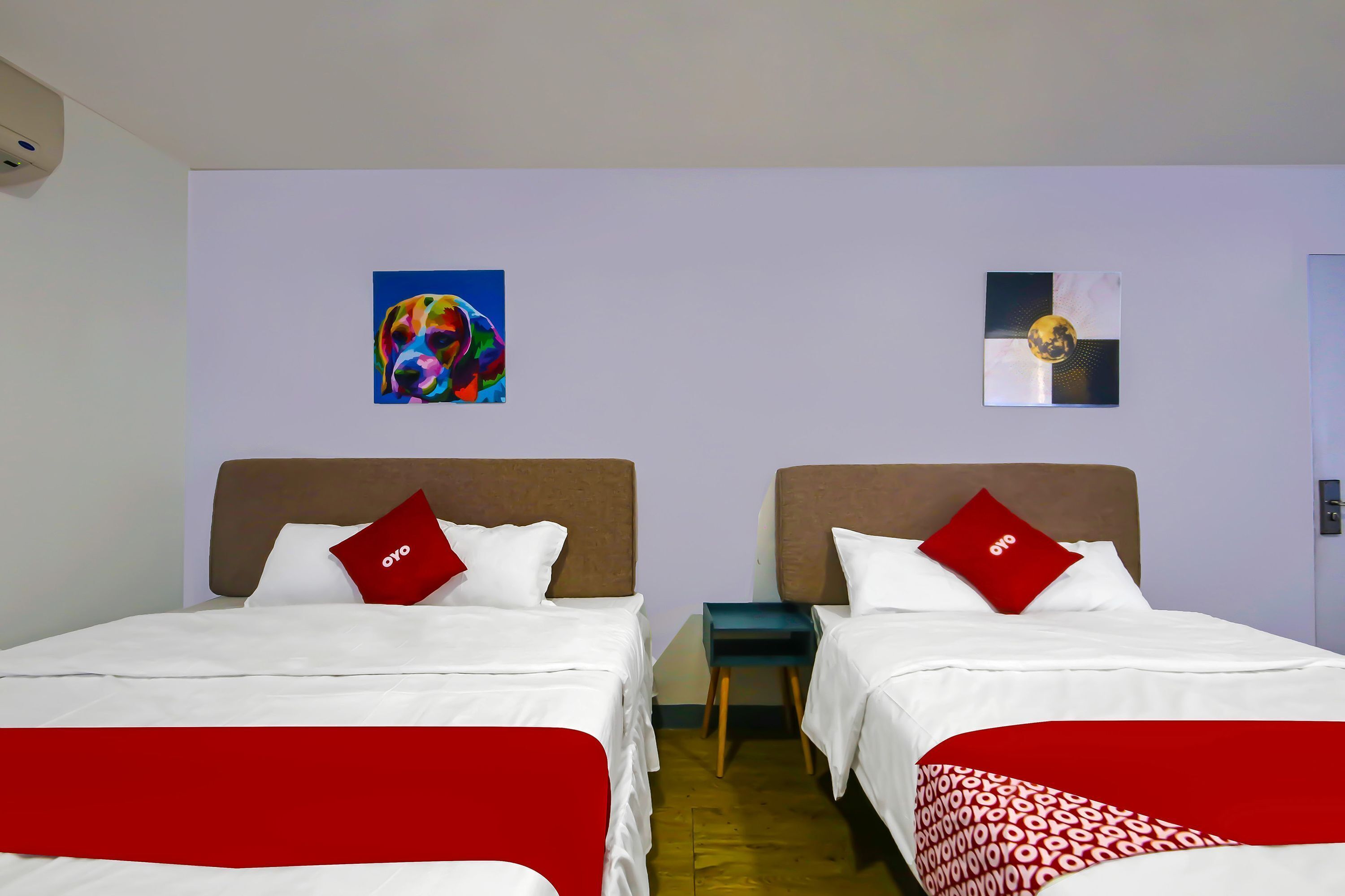 Une chambre d’hôtel avec deux lits, chacun agrémenté d’un oreiller rouge et de draps blancs. Deux œuvres d’art sont accrochées aux murs, une au-dessus de chaque lit.