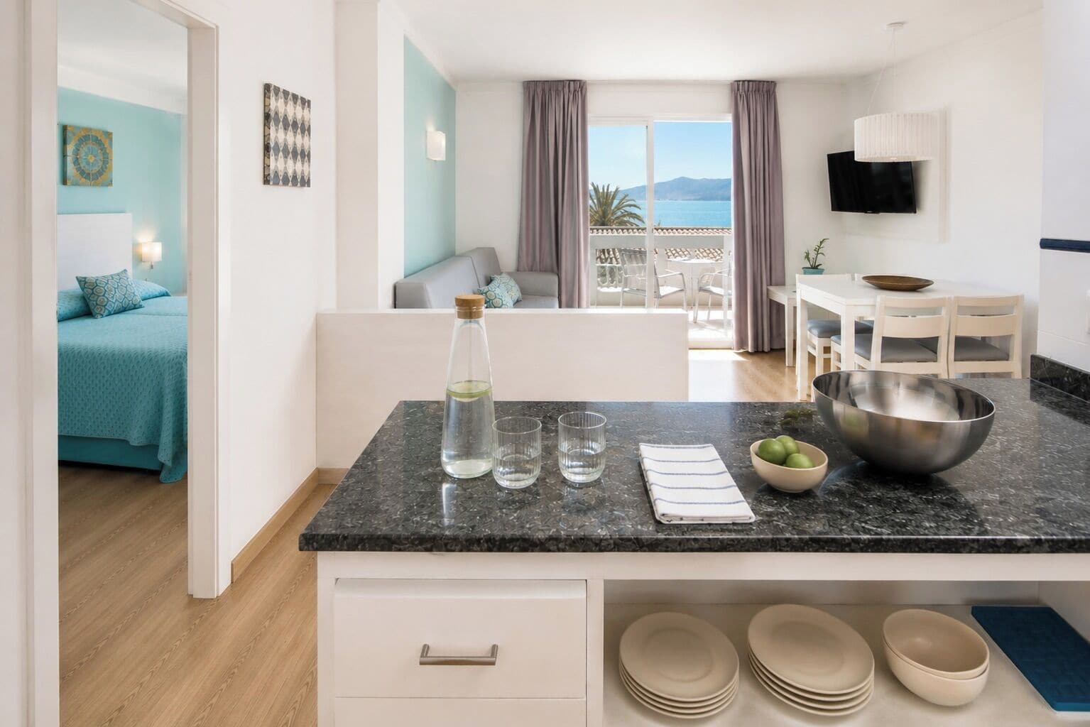 A modern kitchen with a marble countertop, a dining area with a table and chairs, and a bedroom with a bed and a view of the sea.