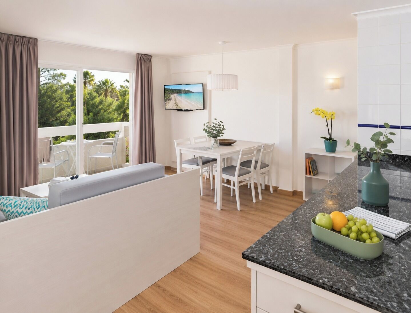A modern kitchen with a dining area, a large window with curtains, a flat-screen TV, and a fruit bowl on the counter.