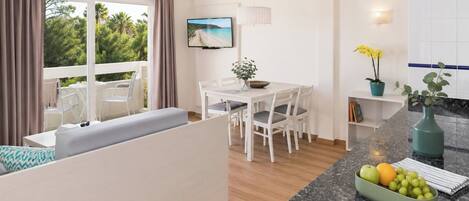 A modern kitchen with a dining area, a large window with curtains, a flat-screen TV, and a fruit bowl on the counter.