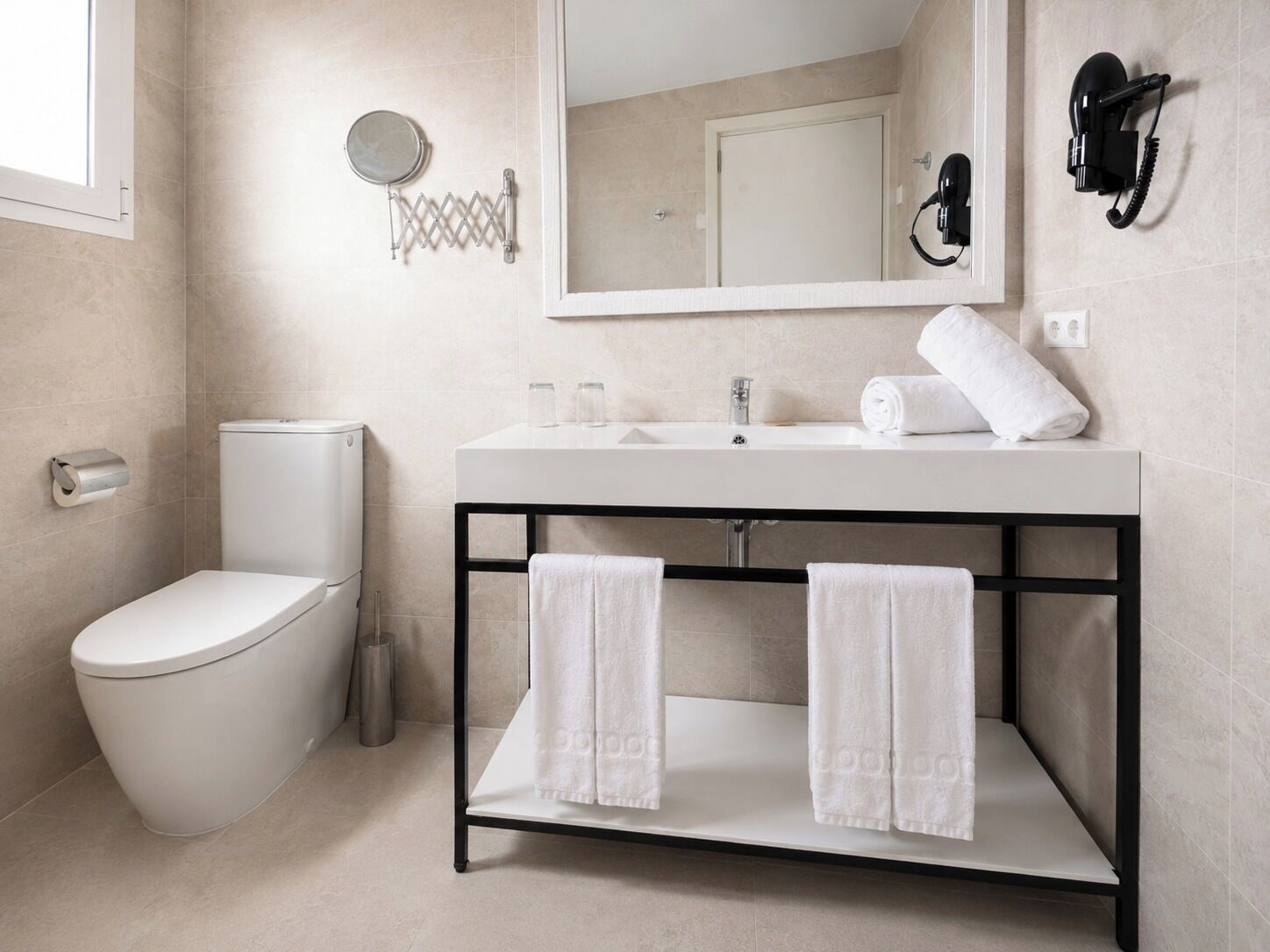 Ein modernes Badezimmer mit Toilette, Waschbecken und Spiegel. Handtücher liegen auf dem Waschbecken und ein Föhn ist an der Wand befestigt.