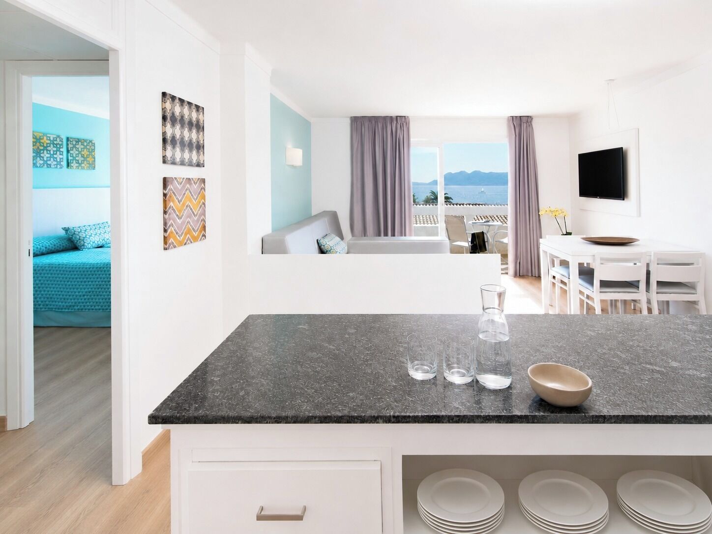 Una cocina moderna con encimera de mármol, zona de comedor con mesa y sillas blancas, y vistas al mar a través de amplios ventanales.