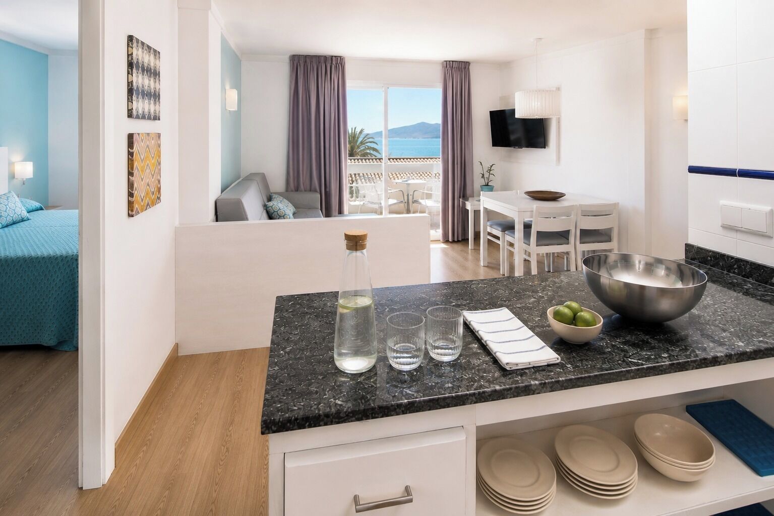 A modern kitchen with a marble countertop, a dining area with a table and chairs, and a bedroom with a bed and a view of the sea.