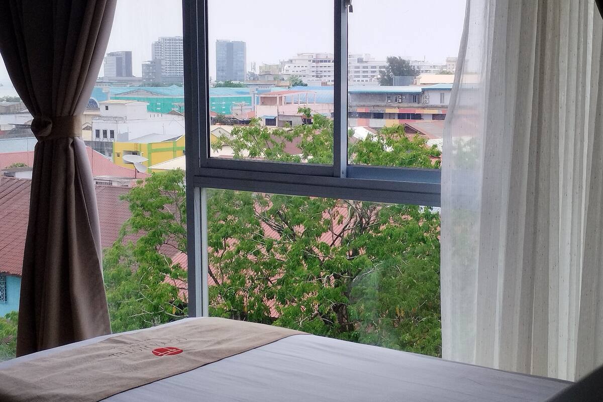 A room with a bed, a window with curtains, and a view of buildings and trees.