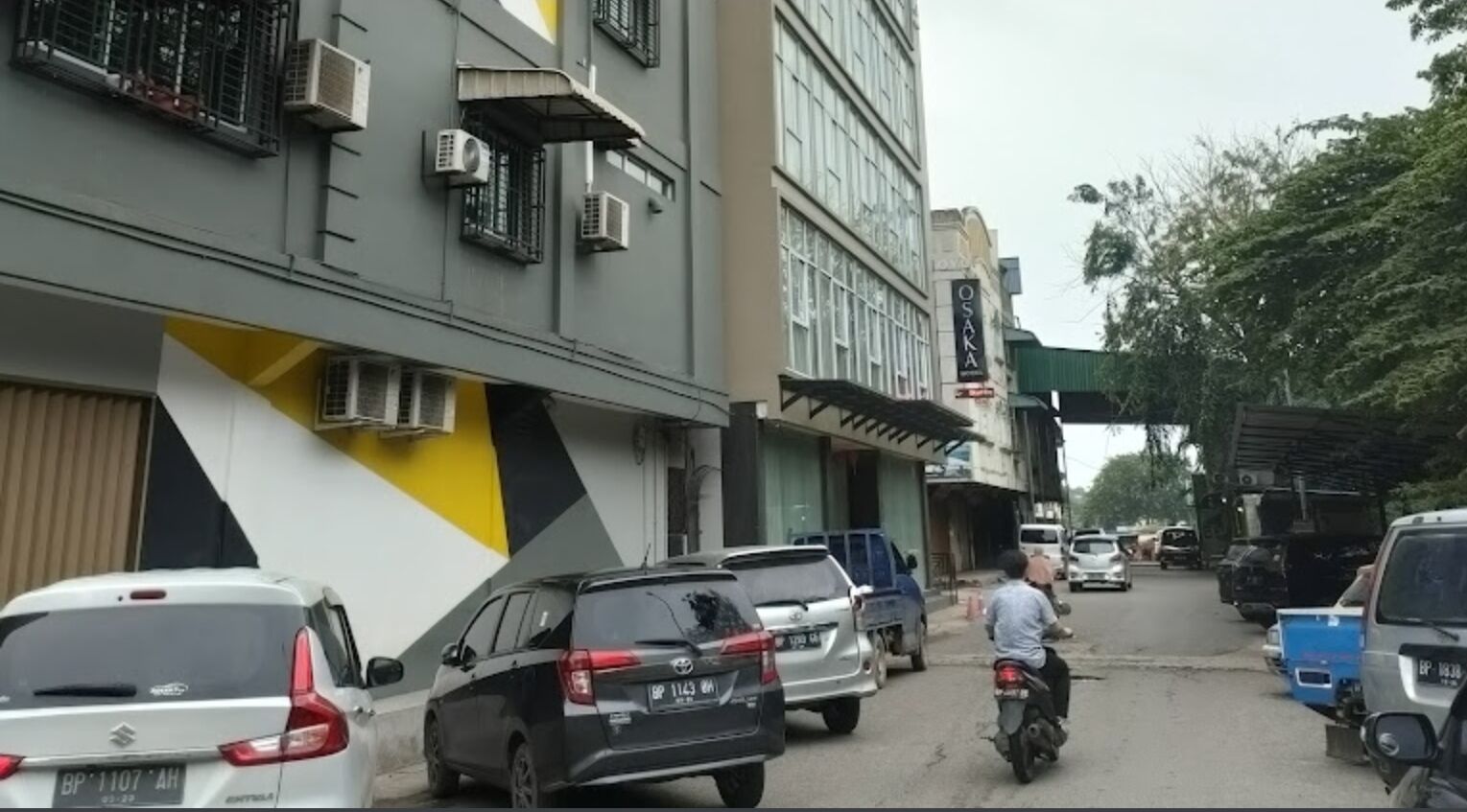 A street with parked cars and a motorcycle. Buildings line the street, and there are trees in the background.