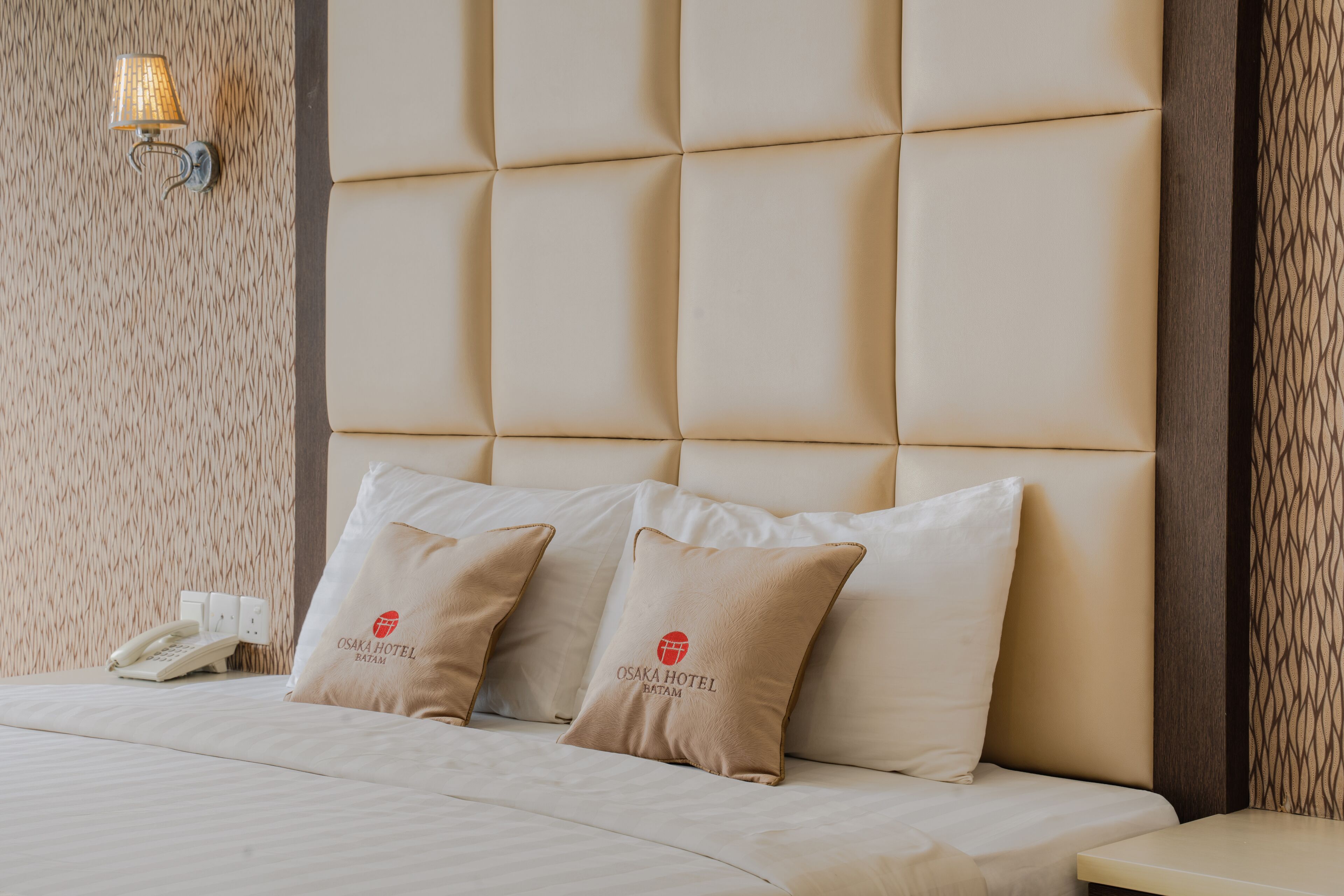 A hotel room with a tufted headboard, two pillows with the hotel logo, a bedside phone, and a textured wall.