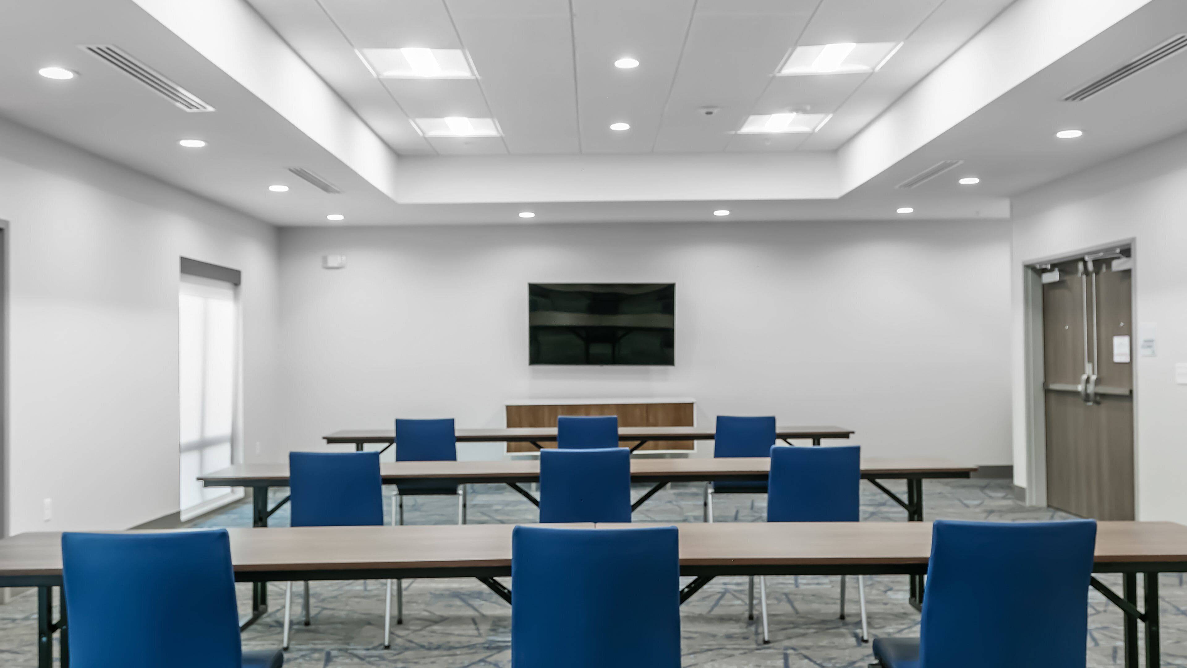 A conference room with a long table, blue chairs, a flat-screen TV on the wall, and a door on the right.
