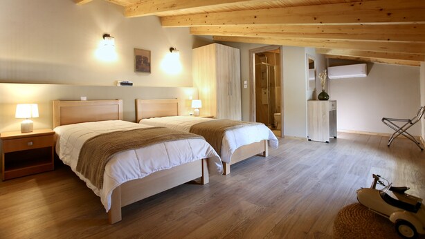 A bedroom with two beds, wooden ceiling beams, a nightstand, a lamp, a chair, and a small table with a vase. - GK Mediterranean Village -AirportShuttle by GK Hospitality Group (Markopoulo Mesogaias)