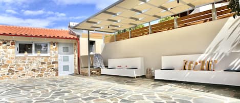 A stone-tiled patio with a white sofa and cushions, a stone building with a red roof, and a pergola with plants.