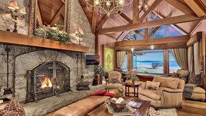 A spacious living room with a stone fireplace, comfortable seating, and a large window overlooking a scenic view. - Lakefront Legacy luxury Lake Tahoe Estate Camp Richardson luxe01 (South Lake Tahoe)