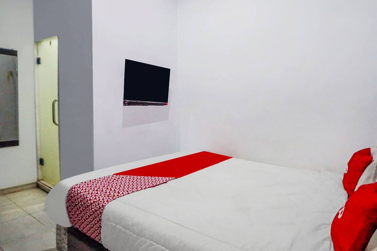 A bedroom with a bed, a red and white patterned blanket, a flat-screen TV mounted on the wall, and a door to the left.