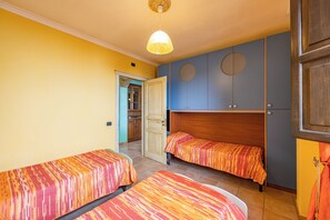A room with two single beds, a wooden headboard, a ceiling light, and a door leading to a kitchen.