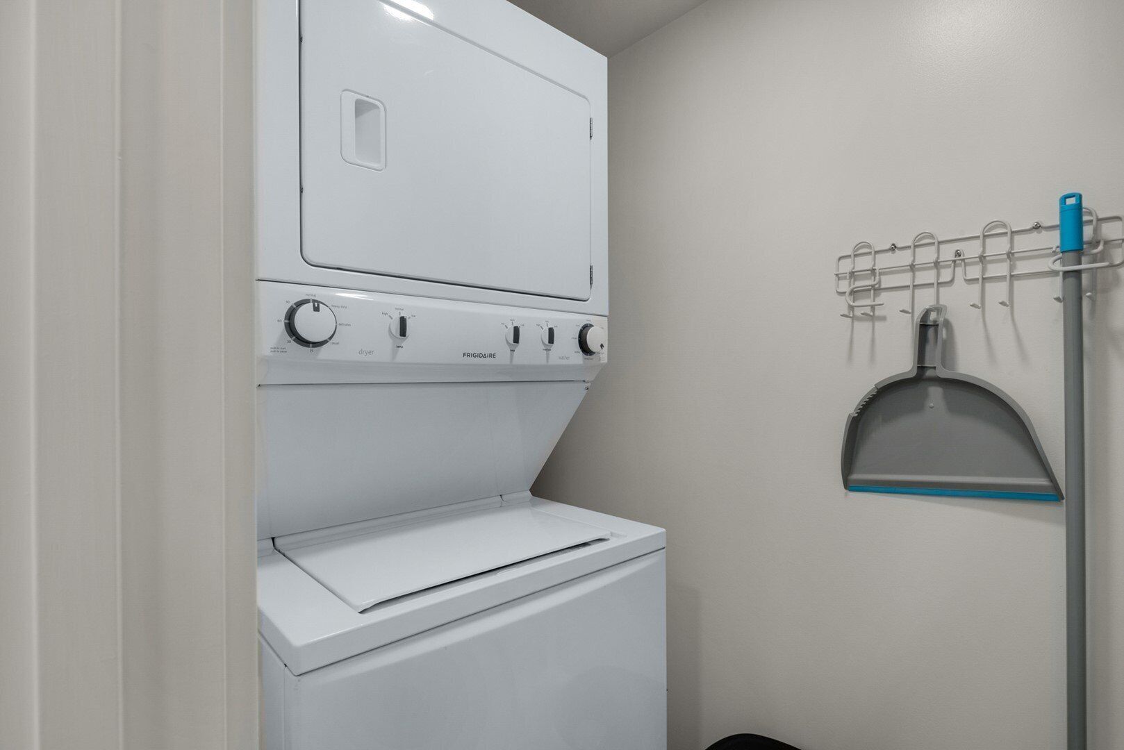 A laundry room with a front-loading washer and dryer, a wall-mounted drying rack, and a broom.