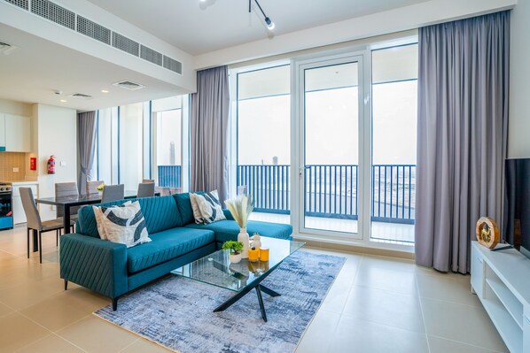 A modern living room with a teal sofa, a glass coffee table, and a large sliding glass door leading to a balcony.