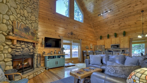 L’intérieur d’une cabane en rondins, avec une cheminée en pierre, des murs en bois, un salon comprenant un canapé et une table basse, et un coin cuisine.