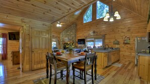 L’intérieur d’un chalet en bois comprend une table à manger, des chaises, une cheminée en pierre, un coin cuisine et une grande fenêtre.