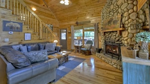 L’intérieur chaleureux d’un chalet avec une cheminée en pierre, un canapé en cuir et un escalier menant à un balcon.