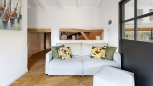 Un salon moderne avec un canapé blanc, des coussins aux motifs géométriques, un plafond avec une poutre en bois et une grande fenêtre.