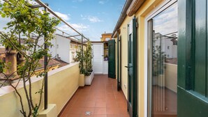 Un étroit couloir extérieur, doté d’un sol carrelé, d’un petit arbre et d’une porte blanche.
