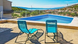 Une piscine avec deux chaises bleues, entourée d’une clôture blanche, donnant sur une ville côtière.