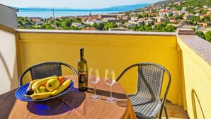 Une table avec une bouteille de vin, deux verres et une assiette de fruits, le tout placé sur un balcon donnant sur une ville côtière.