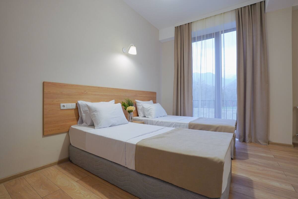 A hotel room with two beds, a wooden headboard, a bedside table with a lamp, and a large window with curtains.