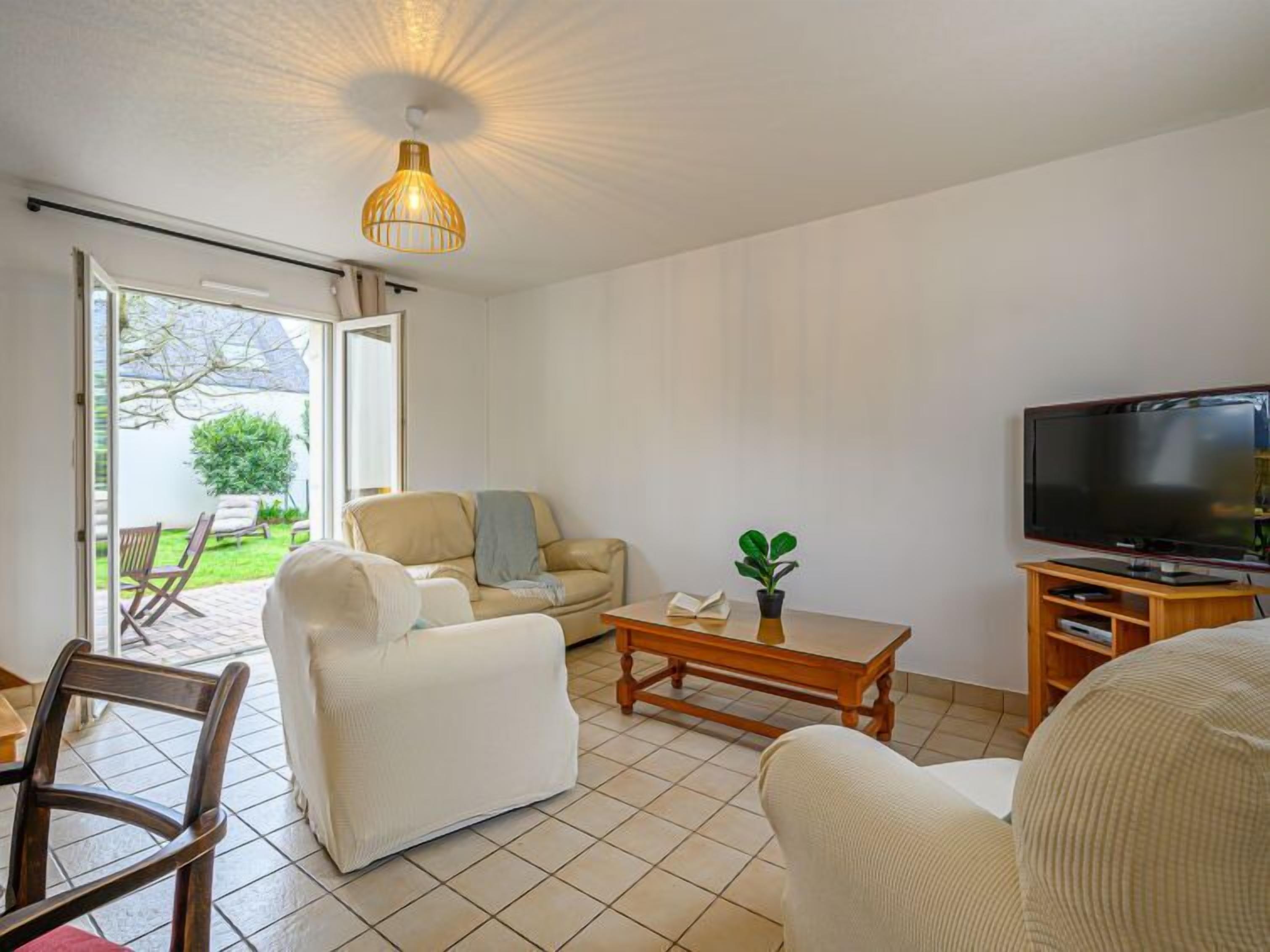 Una sala de estar con sofá, sillón, mesa de centro y televisor. Hay una puerta corrediza de vidrio que da a un área exterior con mesa y sillas de patio.