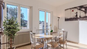 Een eetgedeelte met een ronde tafel en zes stoelen, grote ramen, een radiator en een potplant.