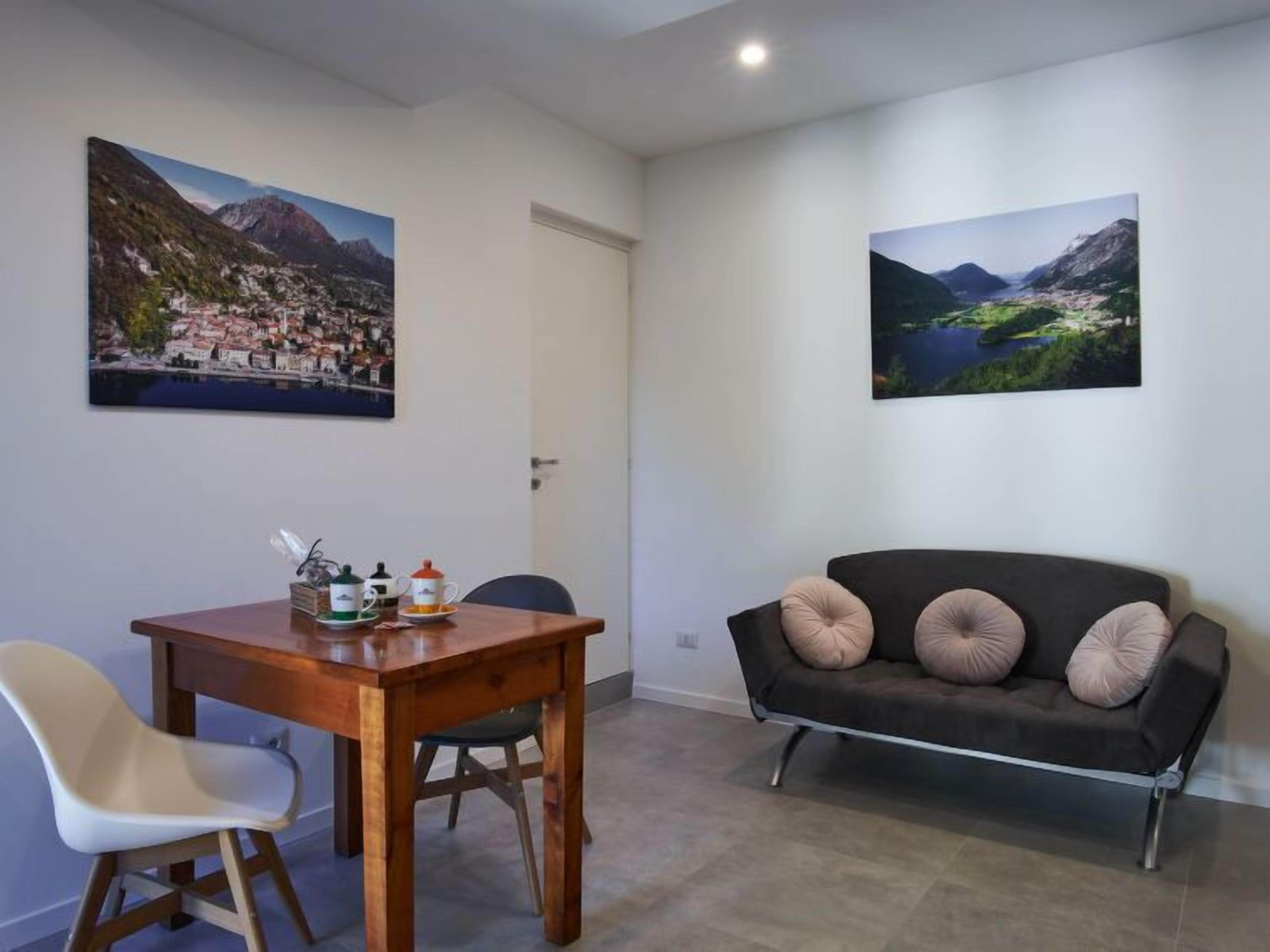 A room with a wooden table, a white chair, a black sofa with cushions, and two landscape paintings on the walls.