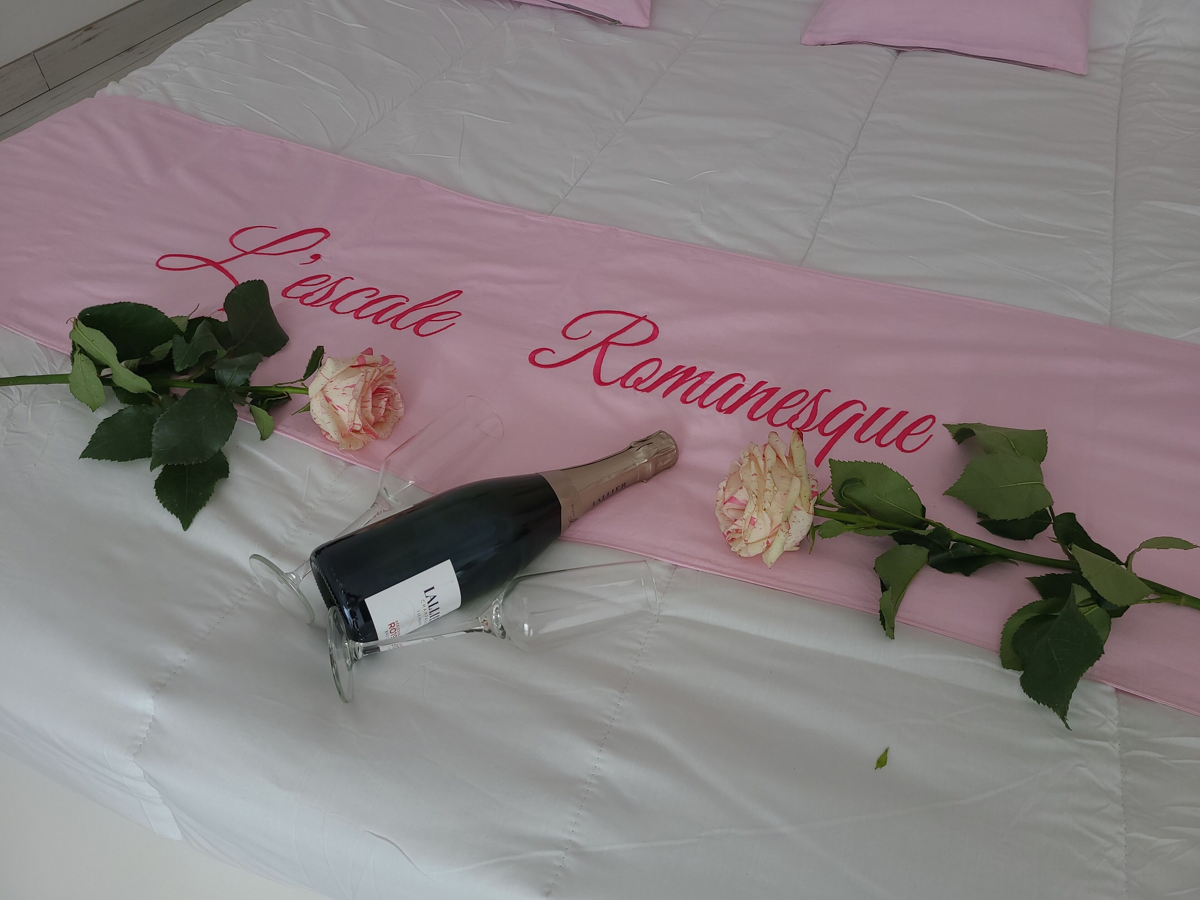 A bed with a pink bedspread that reads "Locale Romaneque" and a bottle of champagne with a glass.