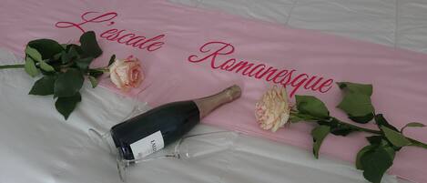 A bed with a pink bedspread that reads "Locale Romaneque" and a bottle of champagne with a glass.