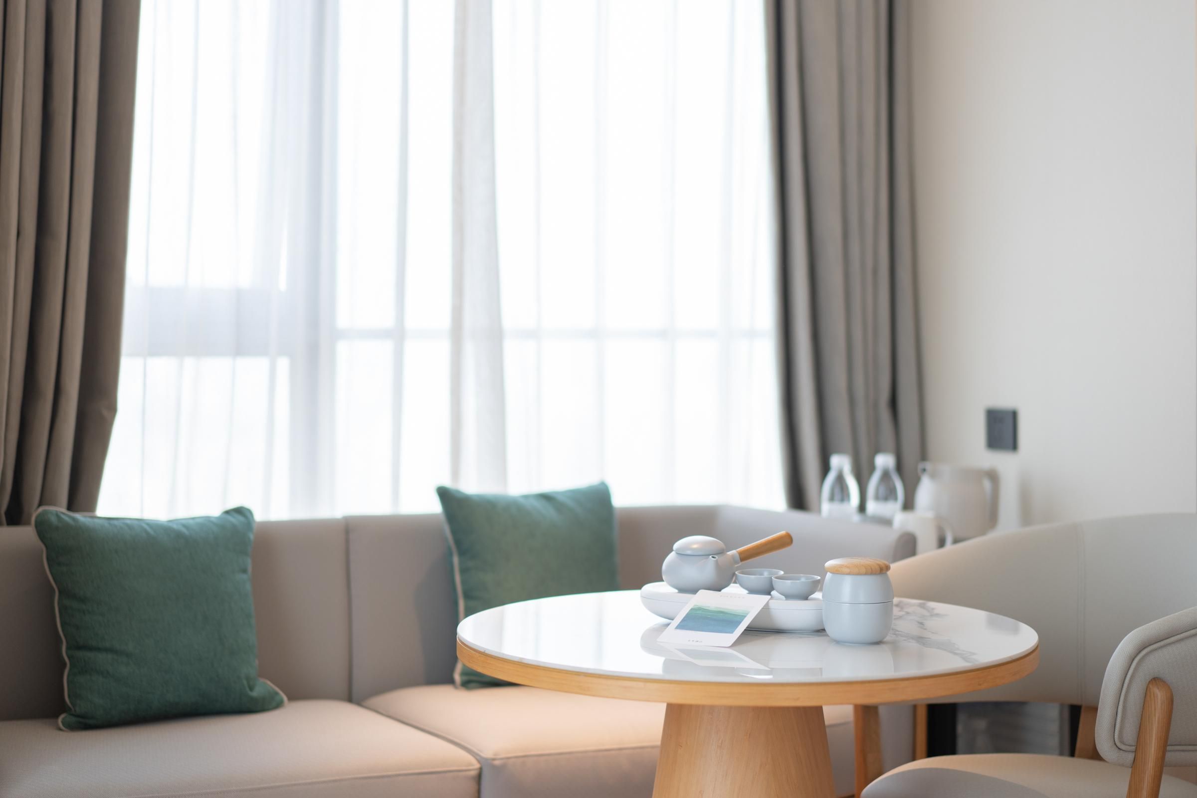 Een moderne woonkamer met een ronde tafel, een beige bank en uitzicht op het groen door grote ramen.