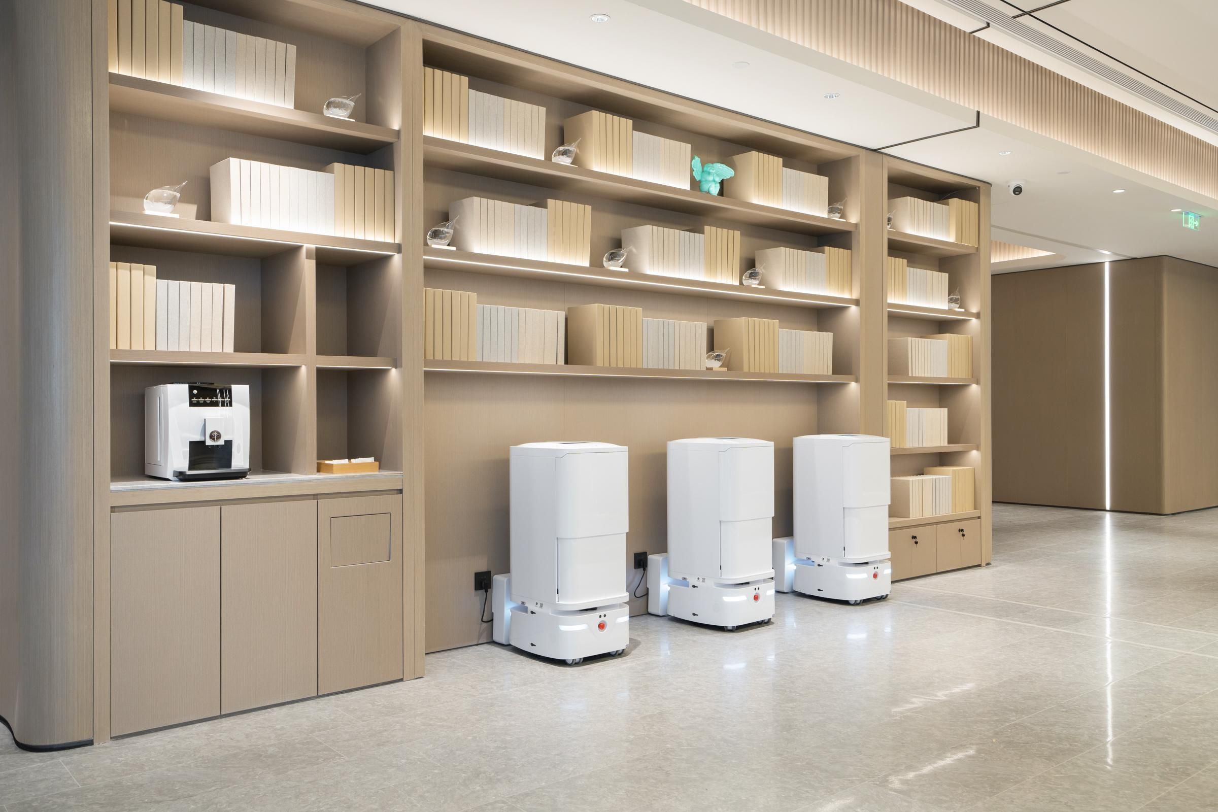 A modern hotel lobby with a clean, minimalist design, featuring a large bookshelf, a coffee machine, and three self-service kiosks.