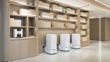 A modern hotel lobby with a clean, minimalist design, featuring a large bookshelf, a coffee machine, and three self-service kiosks.