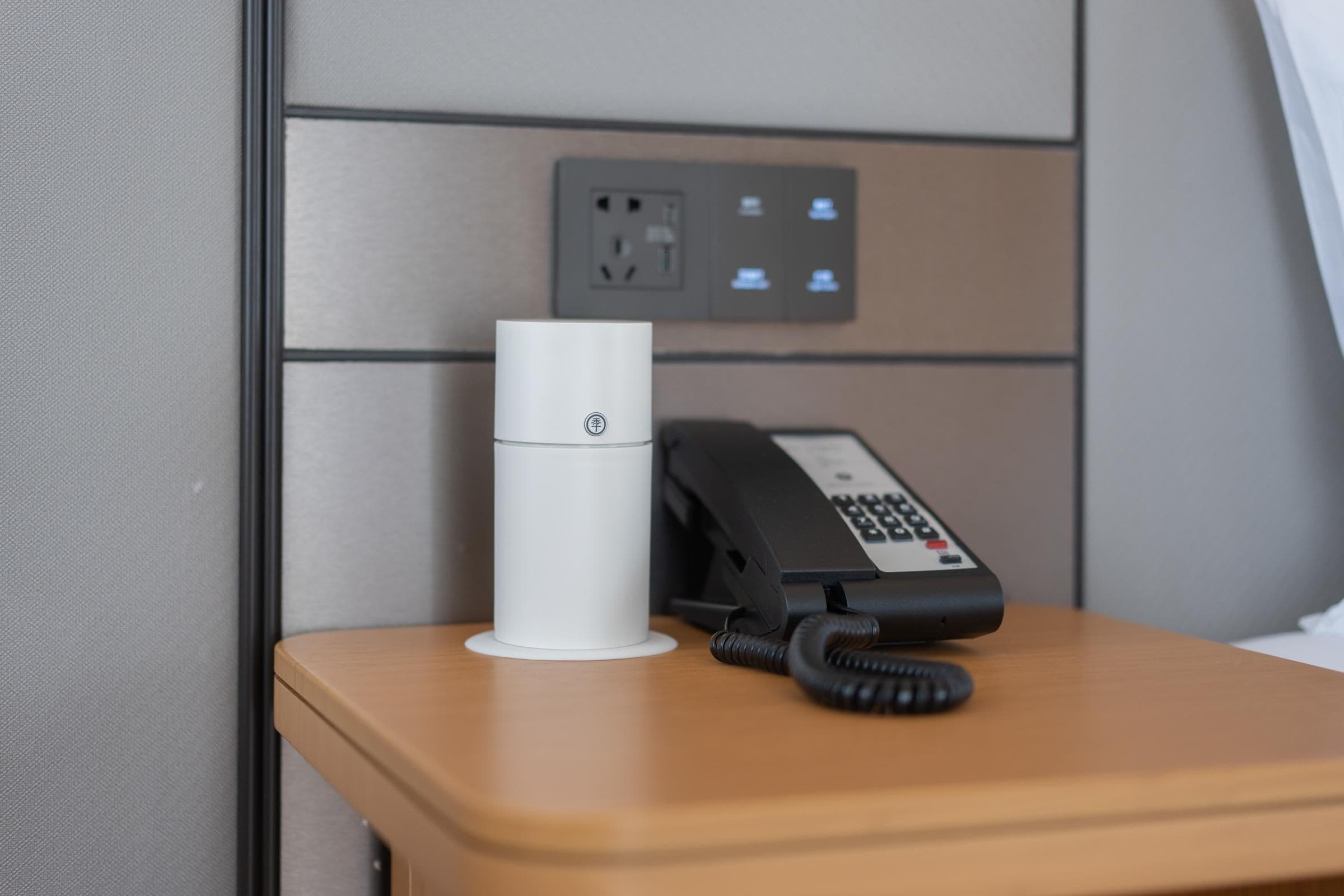 A hotel room with a wooden bedside table, a white cylindrical device, and a black telephone.