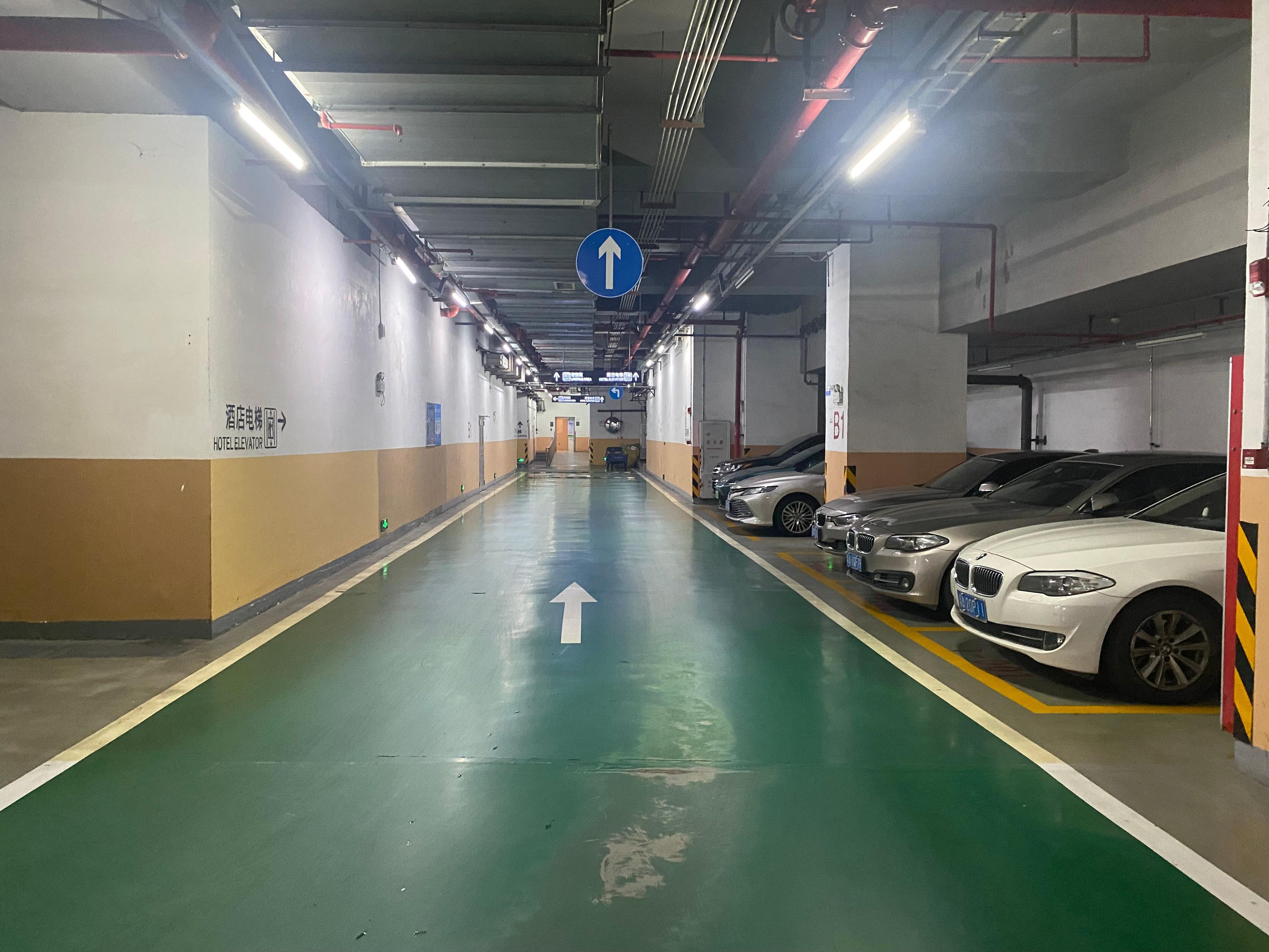 Un estacionamiento de varios niveles con una flecha verde que indica la dirección en el suelo, un letrero de ascensor de hotel y varios automóviles estacionados.