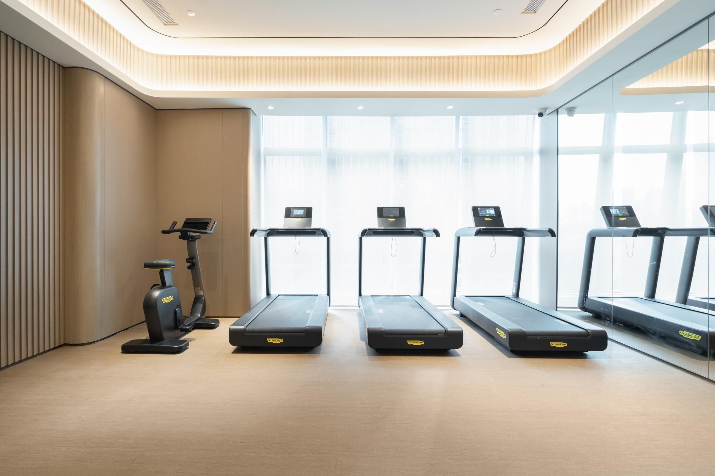 A modern gym with four treadmills aligned in a row, a stationary bike, and a wall-mounted exercise bike.