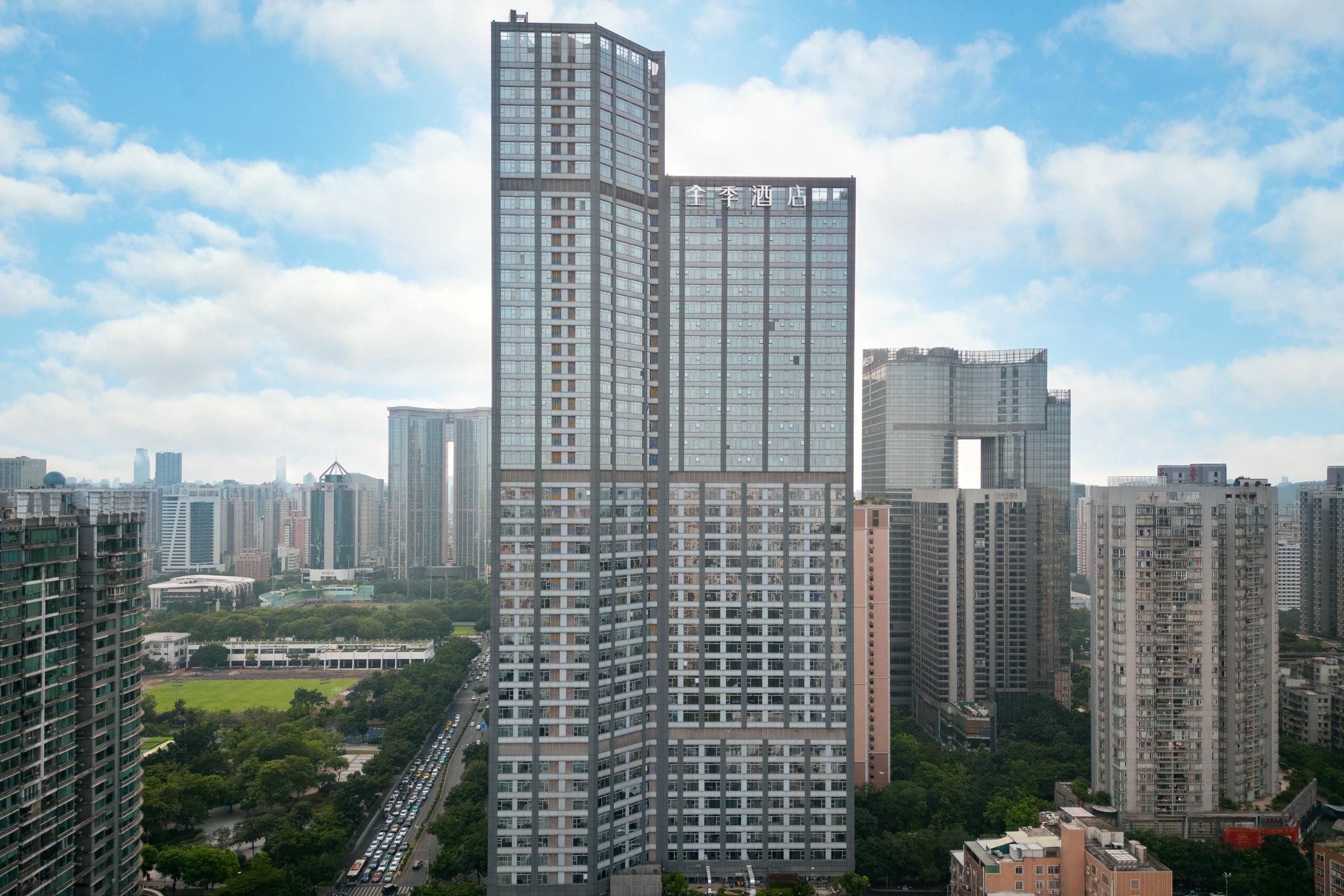 A high-rise hotel with a distinctive angular design, surrounded by other tall buildings and a green park area.