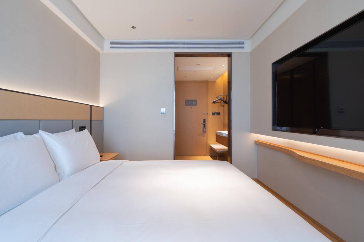 Una habitación de hotel moderna con una cama grande, un televisor montado en la pared y un baño visible a través de una puerta abierta.