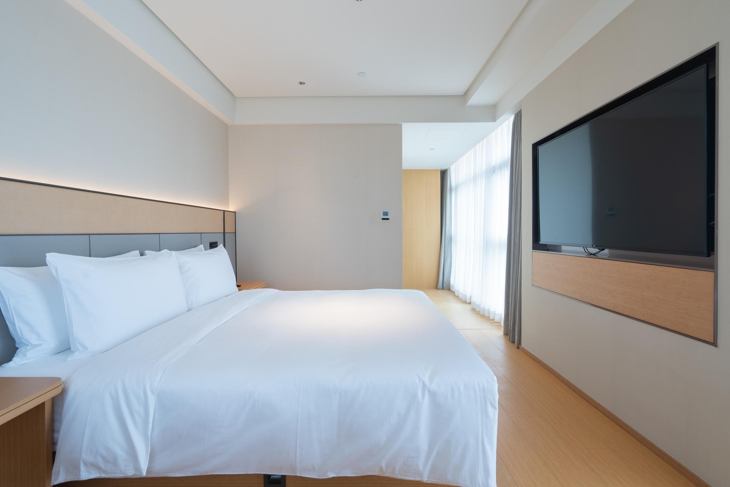 A modern hotel room with a large bed, a flat-screen TV mounted on the wall, and a view of the outdoors through the window.